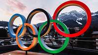 Die olympischen Ringe stehen vor verschneiten Bergen.