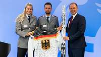 Ein Mann, eine Soldatin und ein Soldat halten ein Paar Skier und ein Trikot in die Kamera.