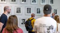 Besucher vor der Porträtgalerie bisheriger Verteidigungsminister und -ministerinnen in Bilderrahmen auf einer weißen Wand