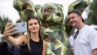 zwei Personen fotografieren sich selbst mit dem Smartphone vor einer Bärenstature in grünem Polygonmuster