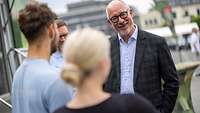 ein älterer Mann mit dunkler Brille und kariertem Anzug lächelt zwei Personen freundlich an 