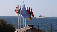 Eine UN-Flagge und Flaggen anderer Staaten wehen im Wind. Dahinter ist Meer und ein Schiff zusehen.