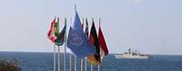 Eine UN-Flagge und Flaggen anderer Staaten wehen im Wind. Dahinter ist Meer und ein Schiff zusehen.