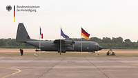 Ein Hercules der deutschen Luftwaffe steht dem Rollfeld. Davor sind drei Flaggen an Masten zu sehen.