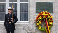 Ein Soldat steht an einer Wand neben einer Gedenkplatte unter der ein Kranz mit Blumen hängt