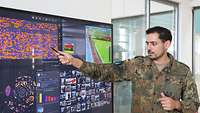 Ein Soldat steht an einem großem Monitor und zeigt mit einem Stift auf einen Teil des Monitors.