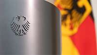 Im Vordergrund steht ein Rednerpult mit dem Bundesadler drauf, unscharf im Hintergrund ist die Deutschlandflagge zu erkennen.