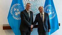 Boris Pistorius und António Guterres stehen vor UN-Flaggen und geben sich die Hand.