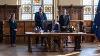 Boris Pistorius sitzt an einem Tisch und unterzeichnet ein Dokument. Hinter ihm stehen zwei Männer.