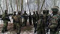 Mehrere Soldaten stehen mit vollständiger militärischer Ausstattung in einem winterlichen Waldgebiet.