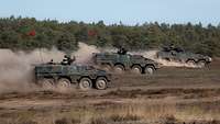 Drei militärische Panzer fahren durch sandiges Gelände