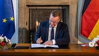 Verteidigungsminister Pistorius sitzt an einem Holztisch und unterzeichnet ein Dokument