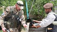Ein deutscher Soldat und ein einheimischer Mann stehen am Brunnen und testen Wasser